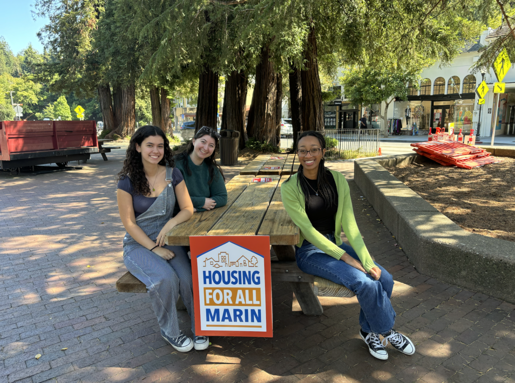 Volunteers sit at a picnic table with a Housing For All Marin sign